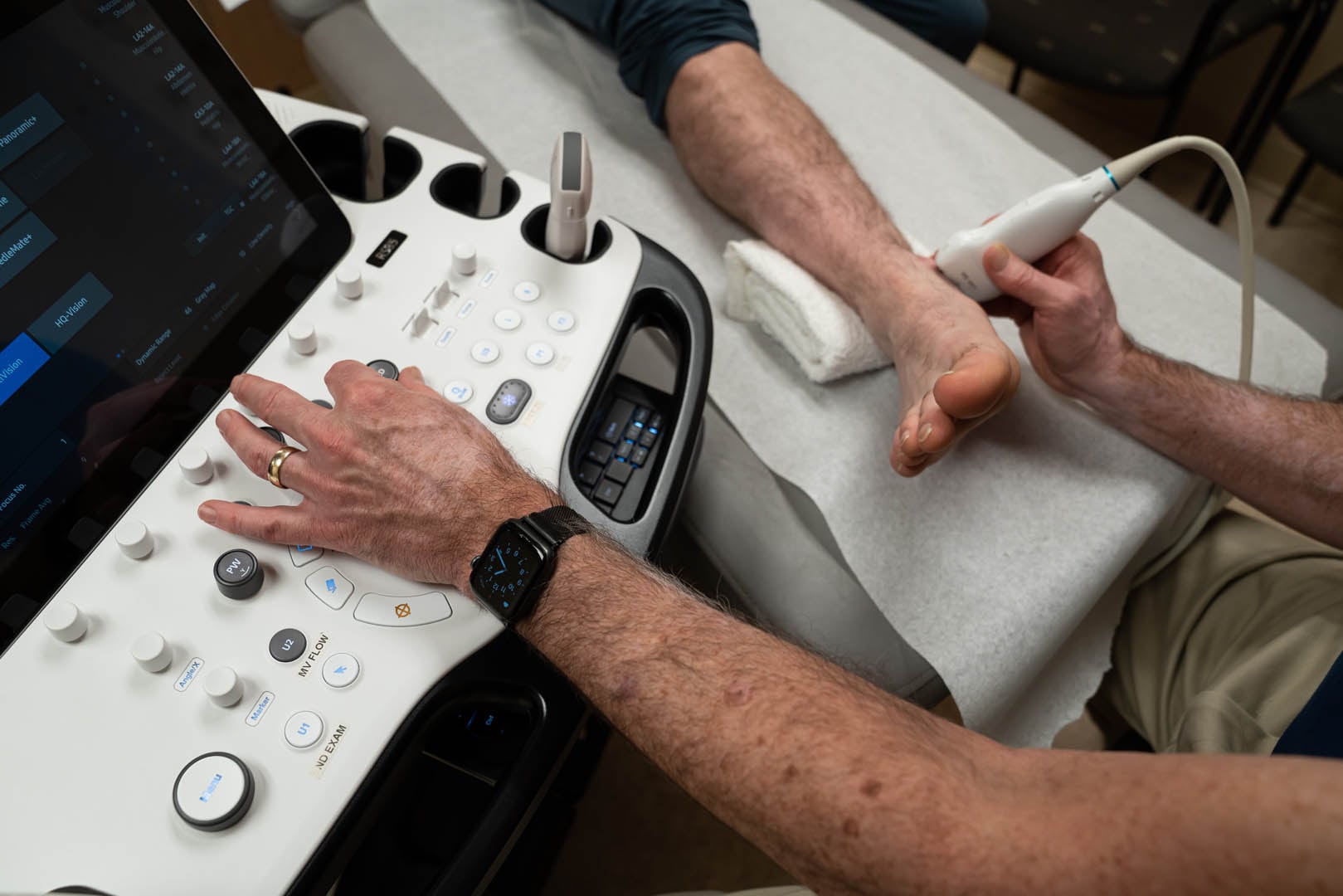 A healthcare professional performs an ultrasound examination on a patient's foot and ankle using a handheld device. The patient's leg rests on a cushioned table, while the professional operates an ultrasound machine with various buttons and controls.