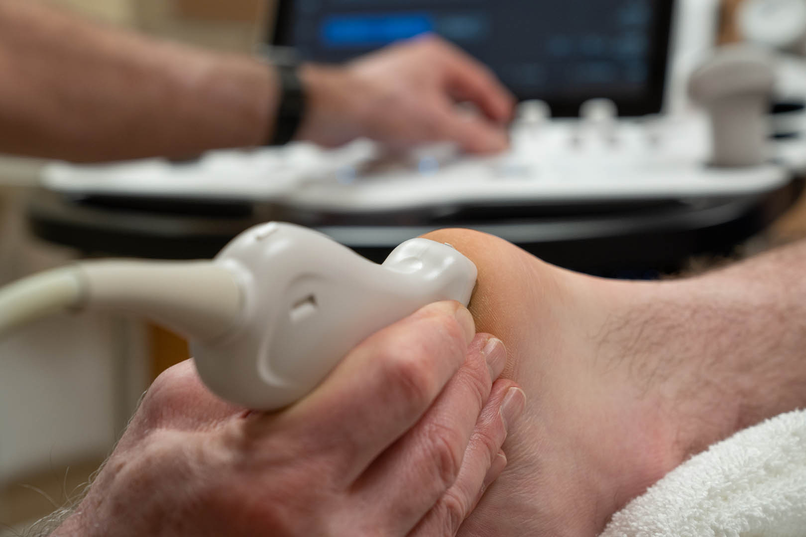 A close-up of a person's heel being examined with an ultrasound device. A medical professional holds the ultrasound wand against the heel, while another hand adjusts settings on an ultrasound machine in the background.