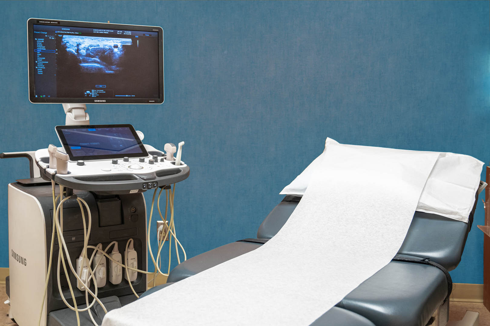 An ultrasound machine is positioned next to a medical examination table covered with a white paper sheet. The machine's screen displays an ultrasound scan, and the room features light blue walls. Various probes and devices are attached to the ultrasound machine.