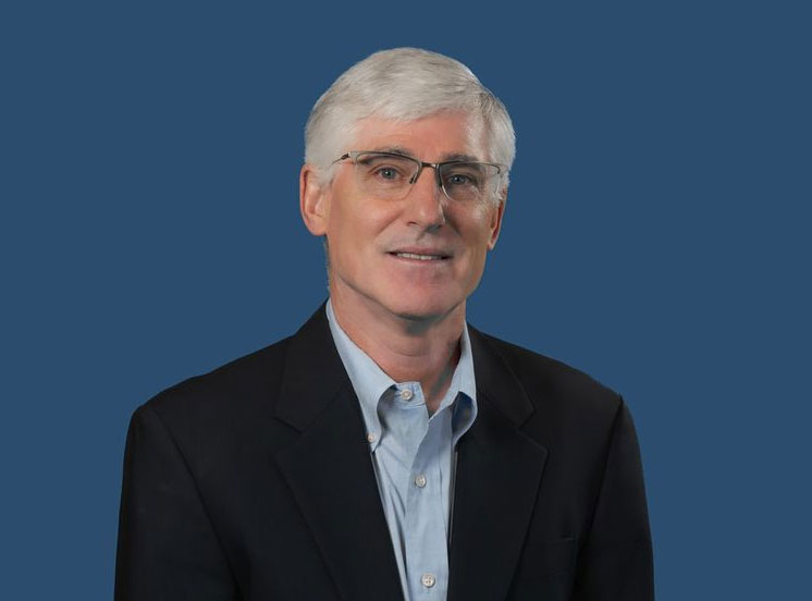 A man with short white hair, wearing glasses, a light blue shirt, and a black blazer poses against a solid blue background. He is smiling slightly.