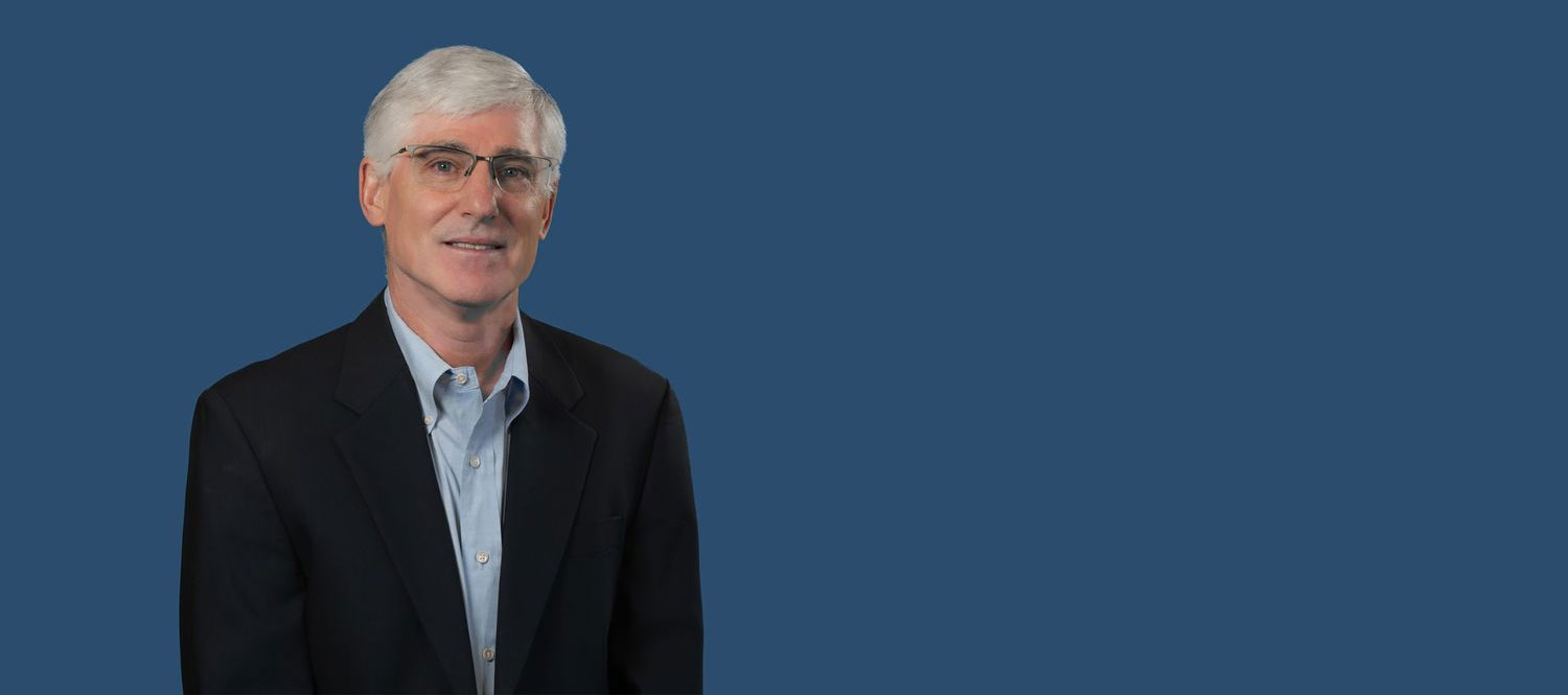 An older man with white hair and glasses is wearing a black suit jacket over a light blue dress shirt. He is smiling slightly and standing against a plain blue background.